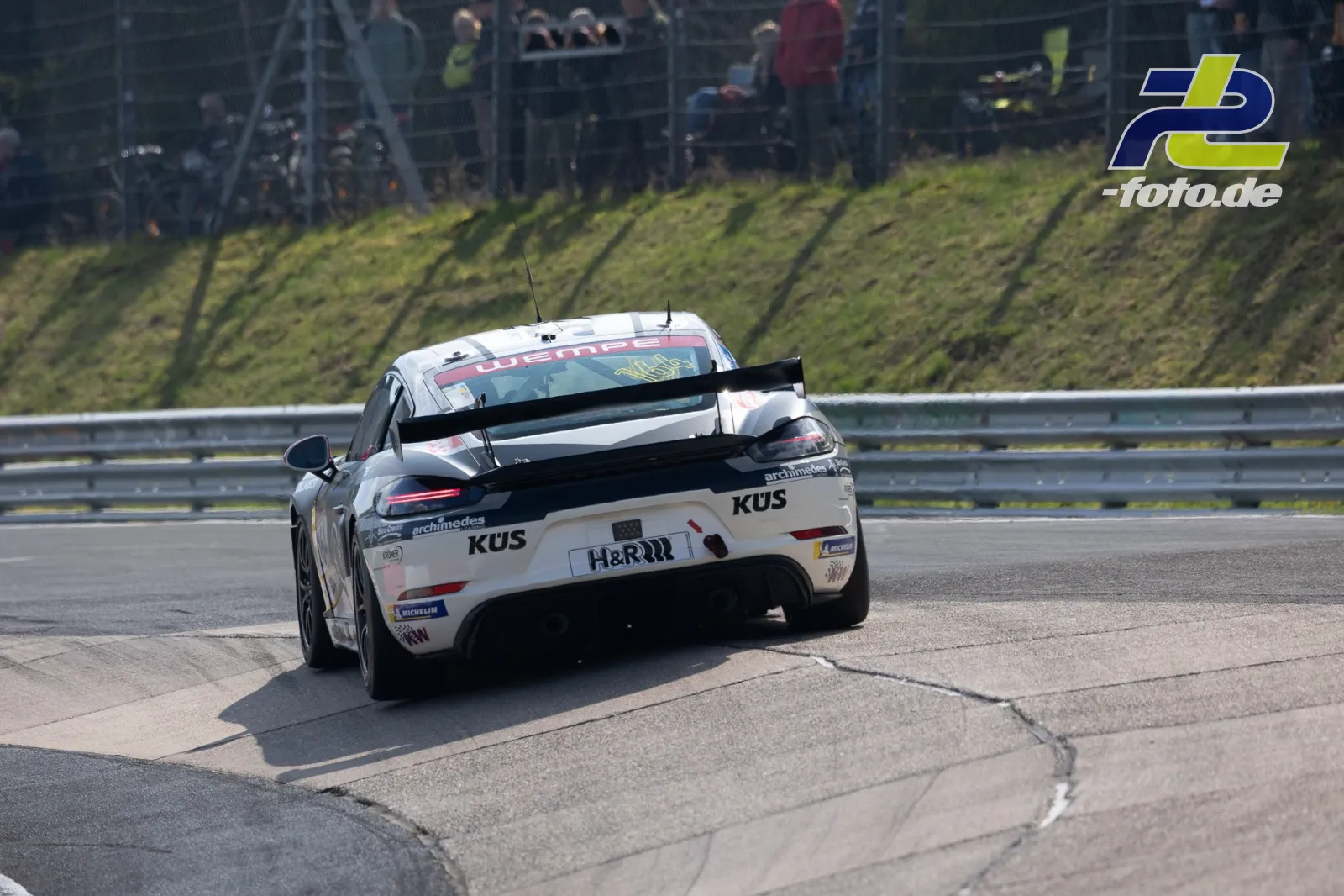 Porsche Cayman mit weißer KUS-Lackierung in der Kurve beim NLS-Rennen auf dem Nürburgring, Heckansicht