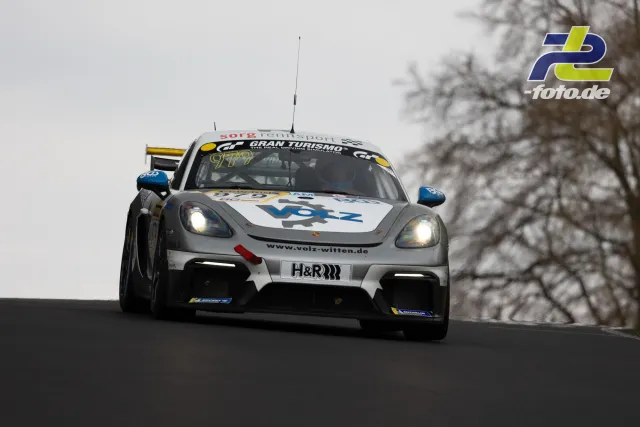 Porsche Cayman GT4 im Sprung über eine Bergkuppe auf der Nordschleife bei bewölktem Wetter, Scheinwerfer eingeschaltet.