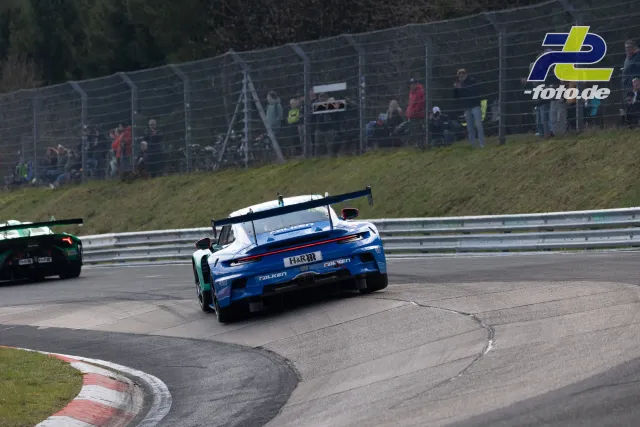 Porsche 911 GT3 R in blau-weißer Lackierung fährt durch eine Kurve auf der Nürburgring-Nordschleife, Zuschauer am Zaun.