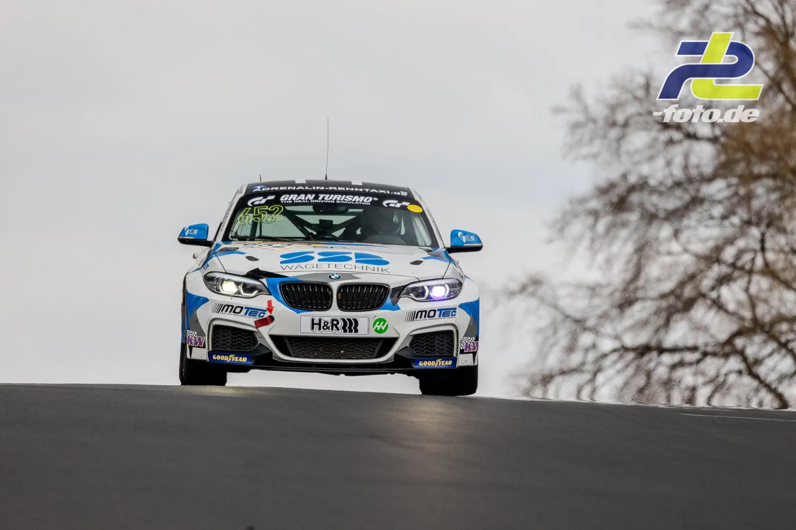BMW M240i Racing frontal über eine Kuppe springend, weiß-blaue Lackierung, NLS Rennen am Nürburgring.