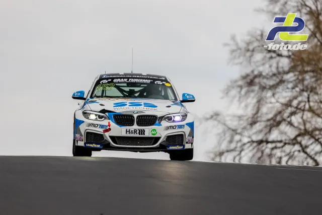 BMW M240i Racing frontal über eine Kuppe springend, weiß-blaue Lackierung, NLS Rennen am Nürburgring.