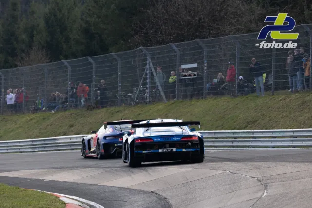 Zwei GT3-Fahrzeuge in der Kurvenverfolgung auf dem Nürburgring, Zuschauer hinter Fangzaun im Hintergrund.