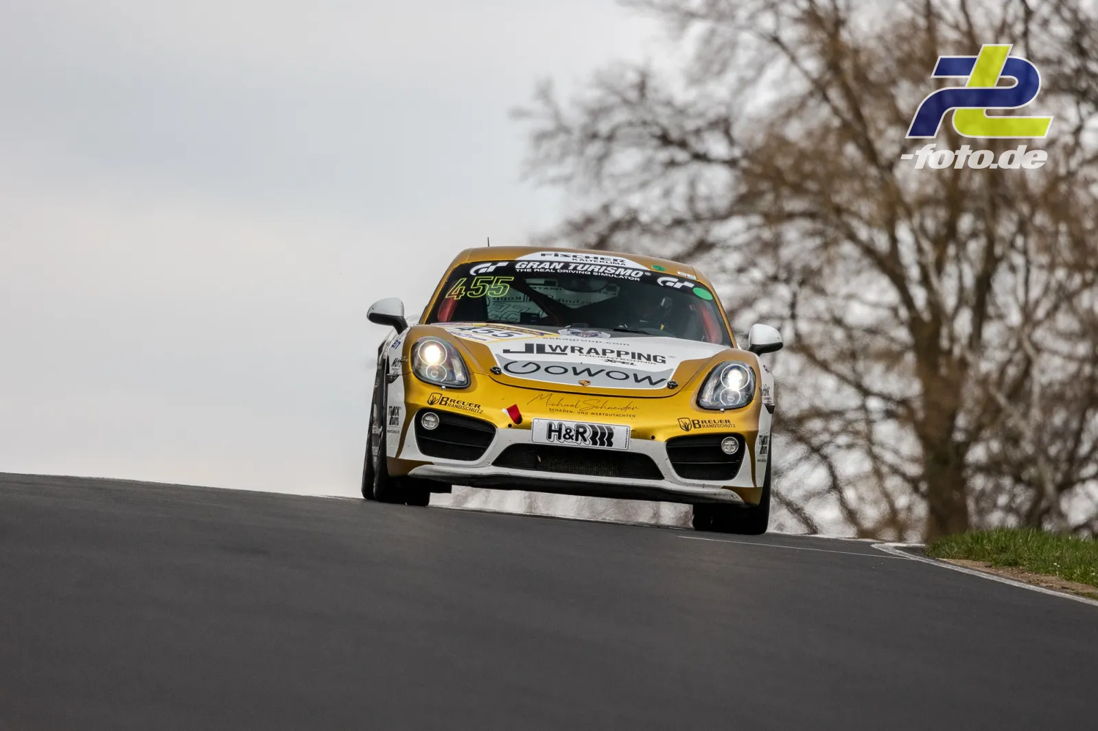 Porsche Cayman mit Startnummer 455 in gelb-weißer Lackierung über die Kuppe am Nürburgring, kahle Bäume im Hintergrund.