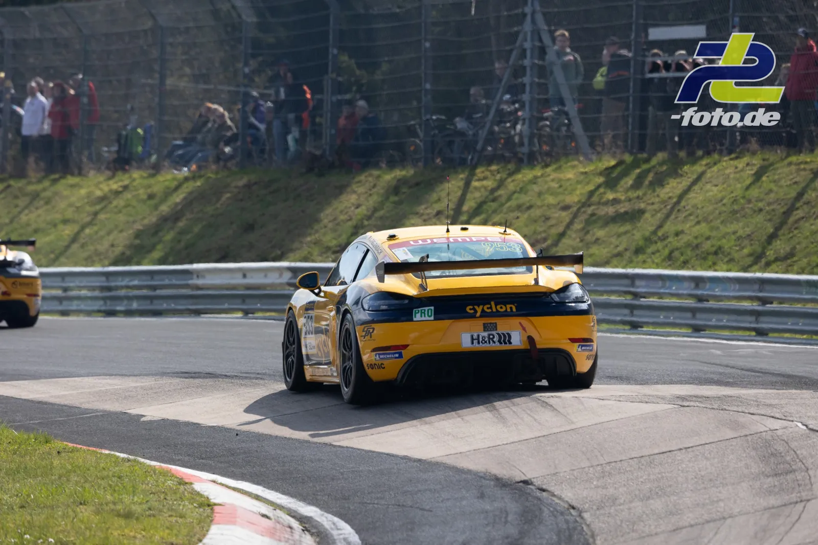 Porsche 718 Cayman GT4 in gelb-schwarzer Lackierung auf der Nordschleife des Nürburgrings, Zuschauer hinter dem Zaun.