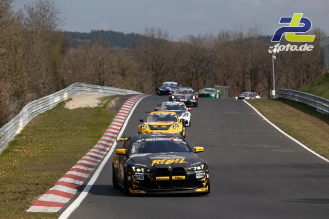 Rennfeld in Nordschleife-Kurve mit BMW in schwarz-gelb gefolgt von gelbem und weiteren Fahrzeugen bei bewölktem Wetter.