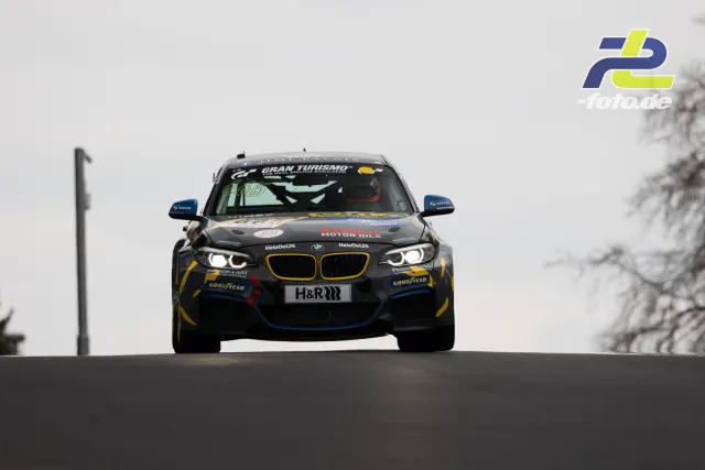 BMW Rennwagen in schwarz-gelb-blauer Lackierung mit Gran Turismo Schriftzug frontal über die Kuppe fahrend bei bewölktem Wetter.