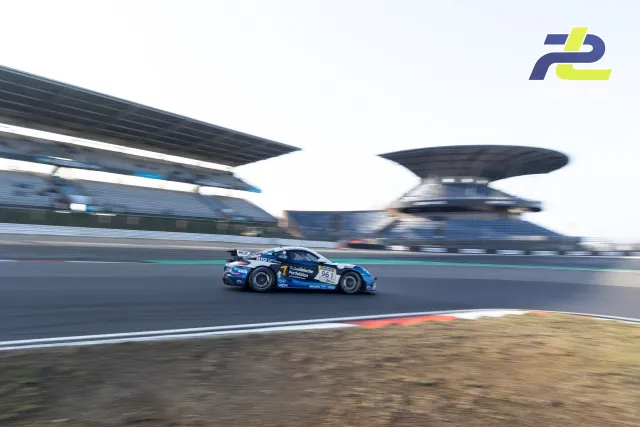 Porsche 961 mit blau-weißer Lackierung im Mitzieher auf der Nürburgring-Grand-Prix-Strecke mit Haupttribüne im Hintergrund.