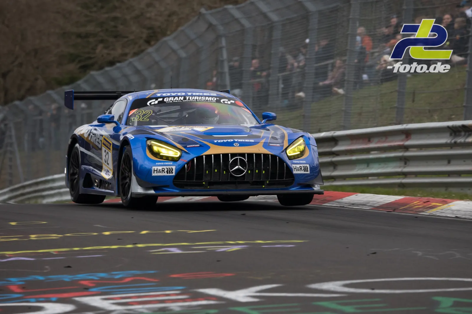 Mercedes-AMG GT3 #32 in Blau-Grau auf der Nürburgring-Nordschleife während des NLS-Rennens mit Zuschauern im Hintergrund.