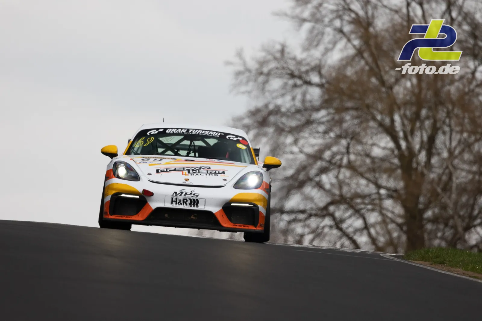 Porsche Cayman GT4 Startnummer 69 in weiß-orange-gelber Lackierung auf der Kuppe am Nürburgring Nordschleife während des Rennens.