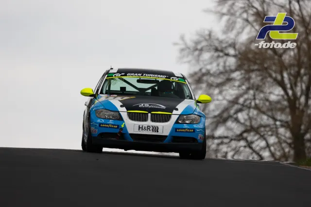 BMW-Rennwagen mit blau-weißer Lackierung und gelben Spiegeln fährt über eine Kuppe auf der Nürburgring-Nordschleife.