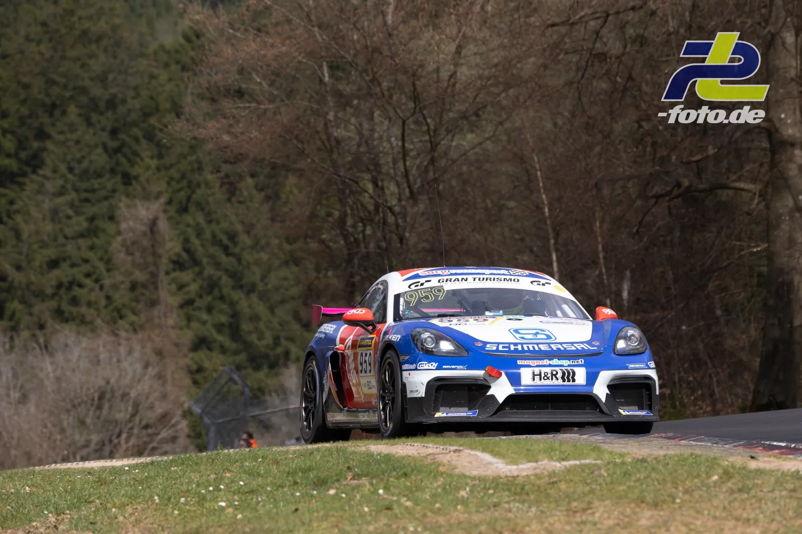 Porsche Cayman GT4 mit blau-weißer Lackierung auf der Nürburgring-Nordschleife bei einer Bergaufpassage, Wald im Hintergrund.