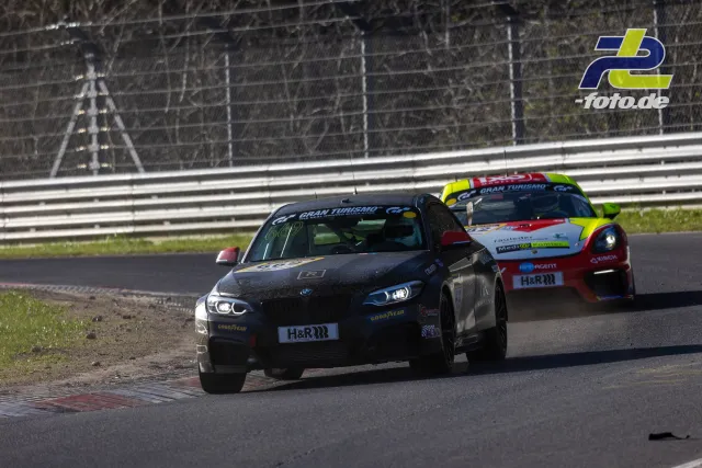 Zwei Rennwagen auf der Nordschleife im Zweikampf, vorderes schwarzes Fahrzeug touchiert Randstein.