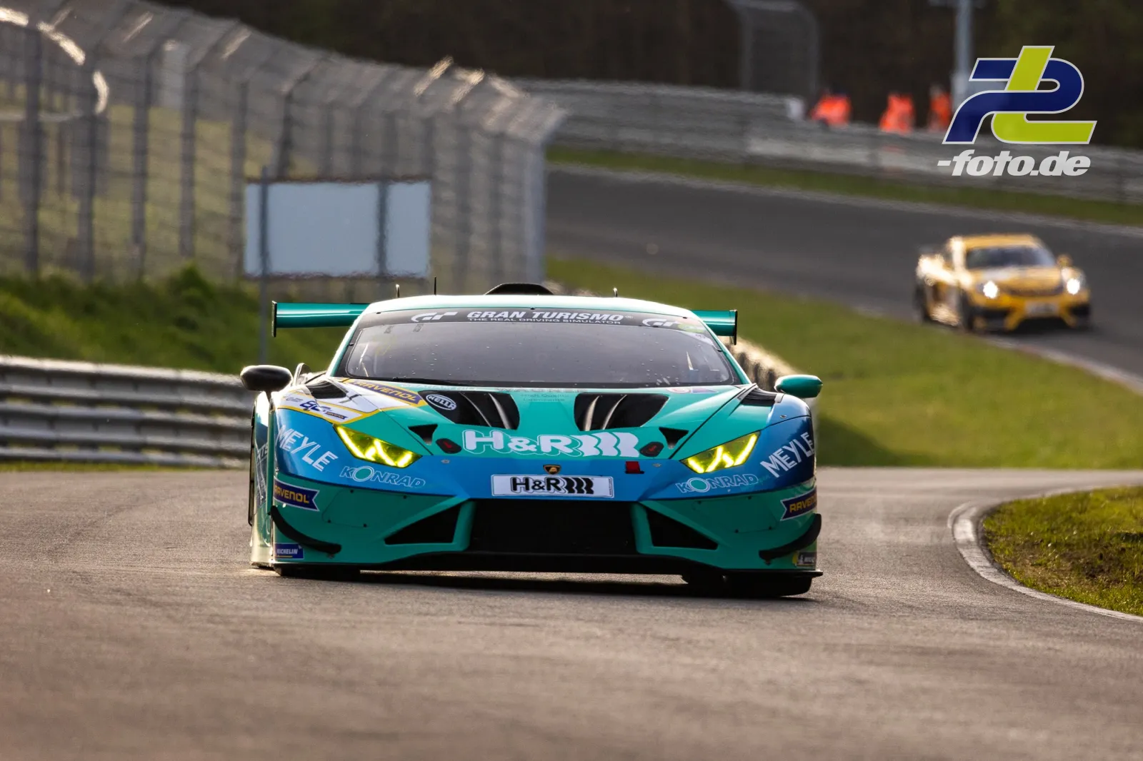 Lamborghini Huracán GT3 in Türkis-Blau auf der Nordschleife, gefolgt von gelbem Porsche im Hintergrund.