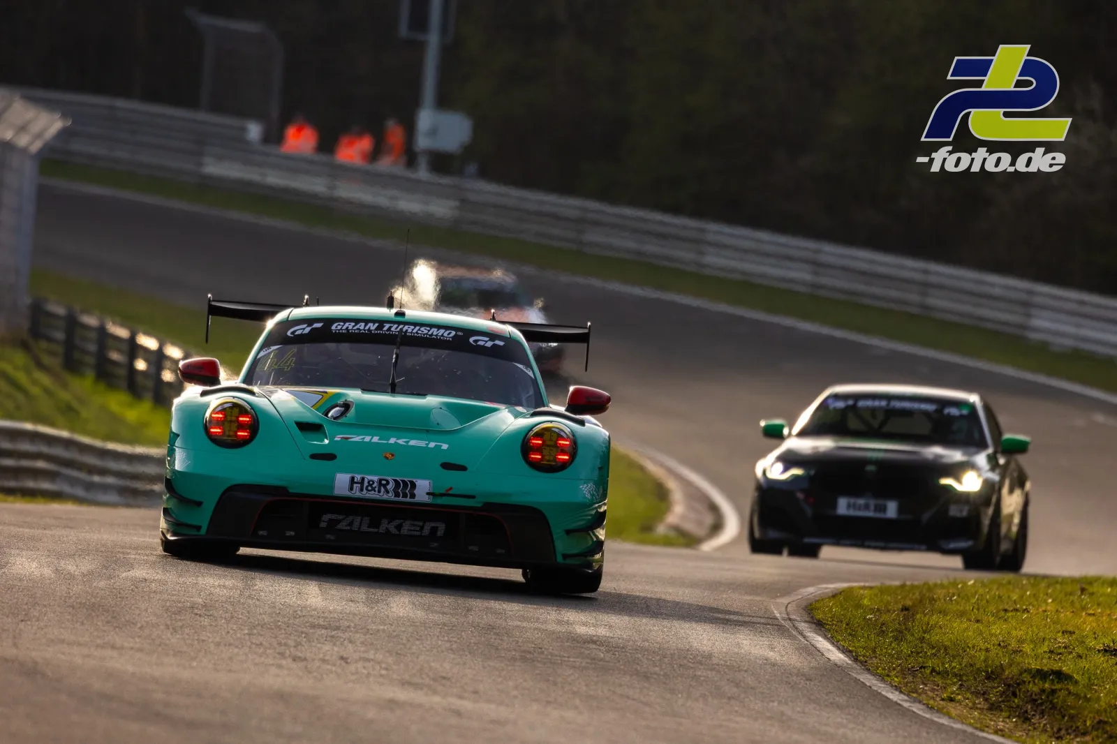 Zwei Rennfahrzeuge in einer Kurve auf dem Nürburgring, vorne ein türkiser Porsche, dahinter ein schwarzer GT-Sportwagen.