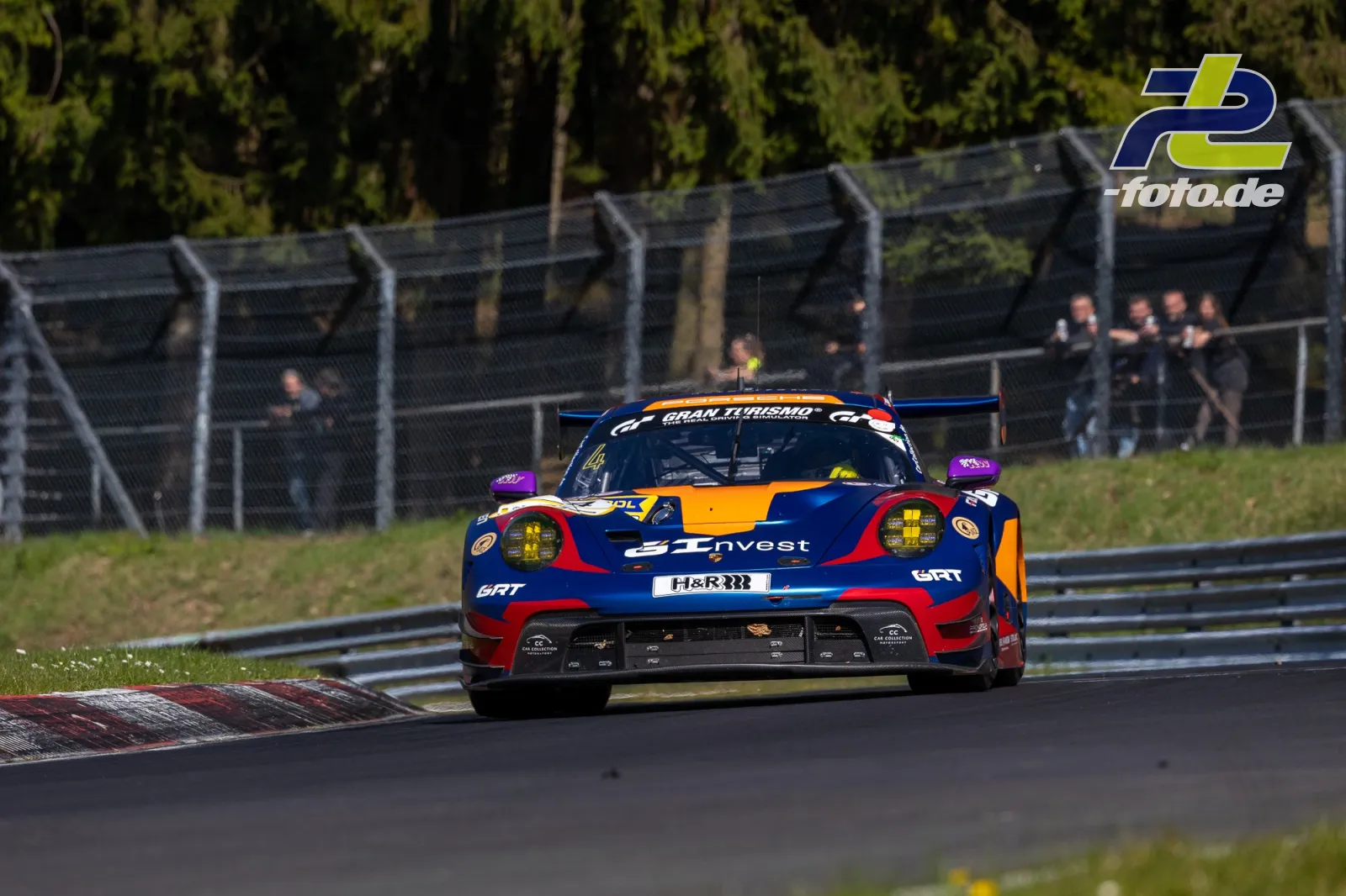Porsche 911 GT3 R in dunkelblau-orangefarbener Lackierung auf der Nordschleife während des NLS-Rennens, Zuschauer hinter Fangzaun.