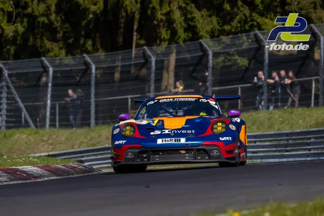Porsche 911 GT3 R in dunkelblau-orangefarbener Lackierung auf der Nordschleife während des NLS-Rennens, Zuschauer hinter Fangzaun.