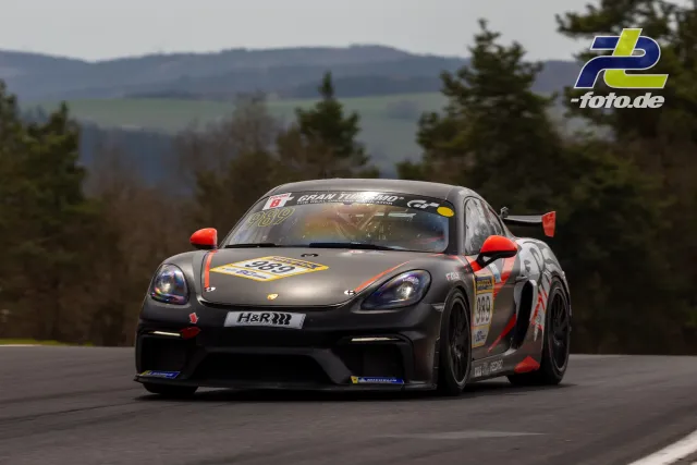 Porsche 718 Cayman GT4 CS in grau-oranger Lackierung bei Kurvenfahrt auf dem Nürburgring, Startnummer 989, Berge im Hintergrund.