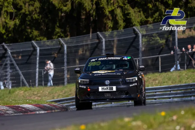 Schwarzer Opel Rennwagen Nr. 380 in Kurvenausfahrt auf dem Nürburgring, Zuschauer hinter Fangzaun, grüne Streckenumgebung.
