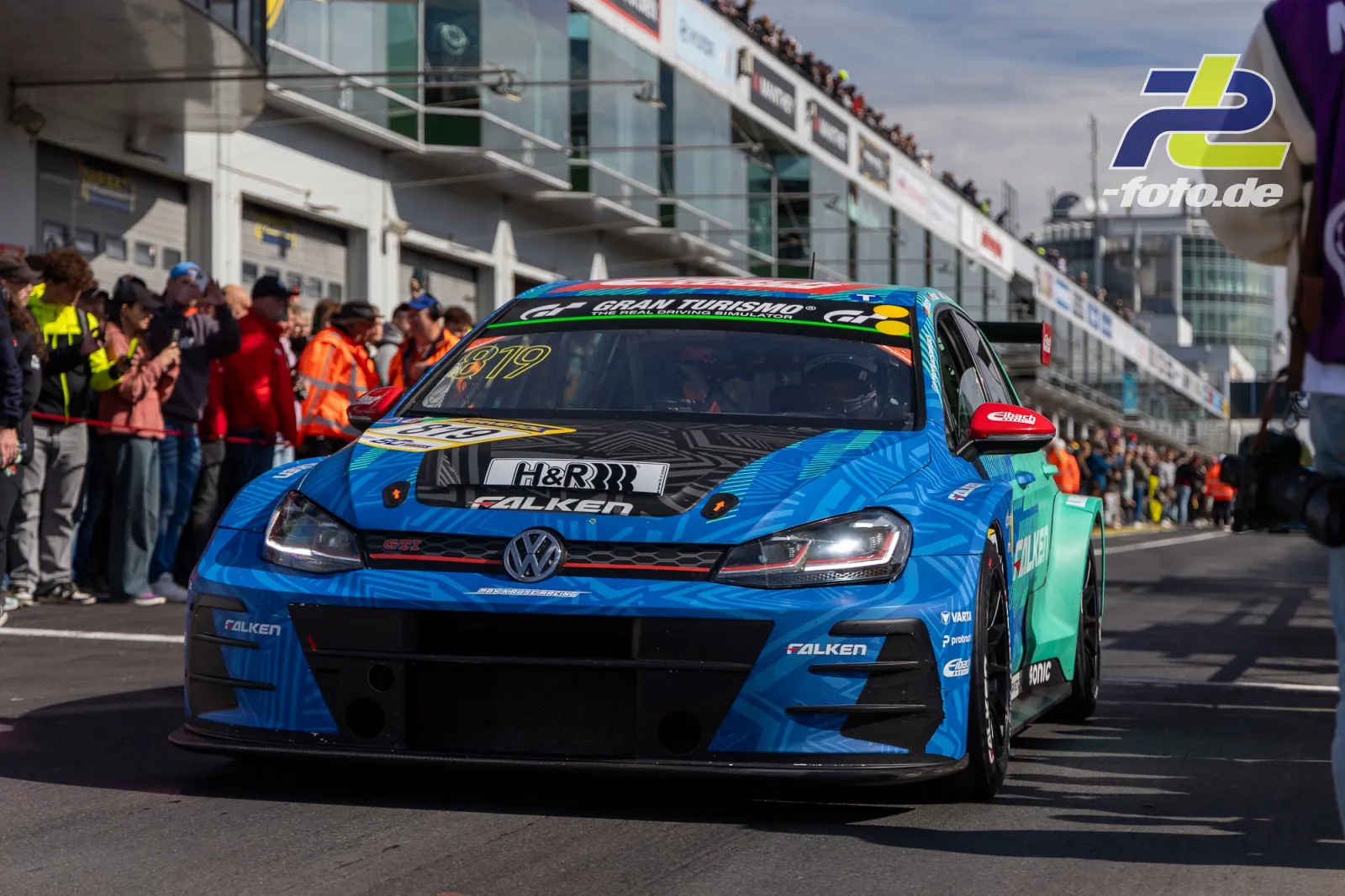 VW Golf GTI TCR mit blau-grüner Falken-Lackierung fährt durch die Boxengasse am Nürburgring, Zuschauer am Rand.