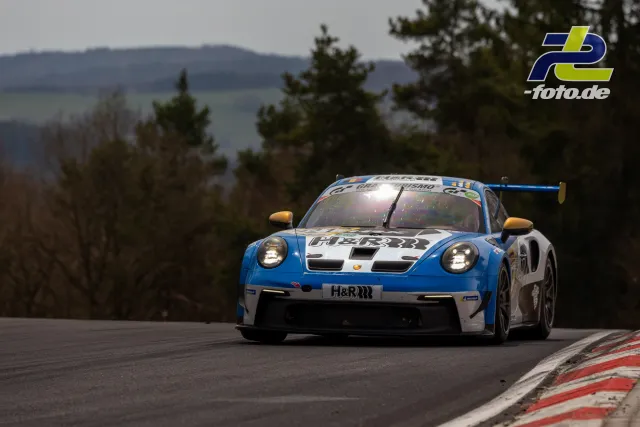 Porsche 911 GT3 R in blau-weißer H&R-Lackierung auf der Nordschleife mit Bergen und Bäumen im Hintergrund.