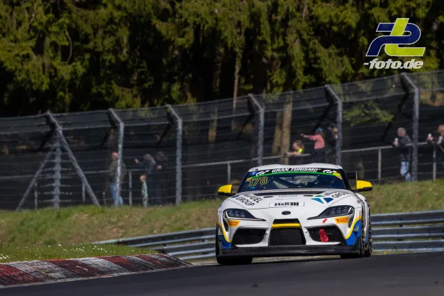 Toyota GR Supra Nummer 178 in weiß-gelb-blauer Lackierung auf der Nordschleife beim NLS-Rennen. Zuschauer hinter dem Fangzaun.