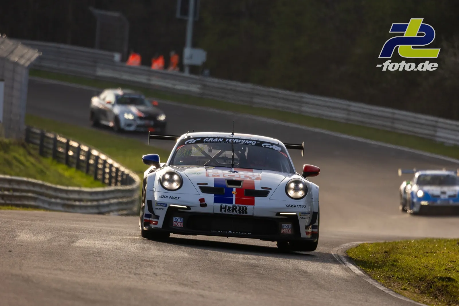 Weißer Porsche 911 GT3 R mit bunter Pixelgrafik in Kurve auf der Nordschleife, mehrere Verfolger im Hintergrund.