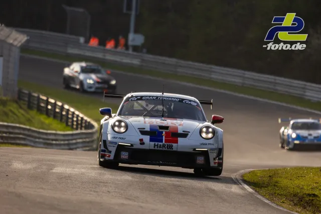 Weißer Porsche 911 GT3 R mit bunter Pixelgrafik in Kurve auf der Nordschleife, mehrere Verfolger im Hintergrund.