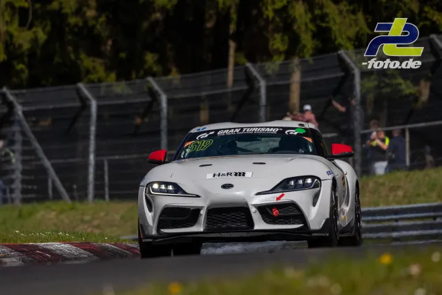 Weißer Toyota GR Supra Nr. 512 mit roten Spiegeln in einer Kurve während des NLS-Rennens am Nürburgring, Zuschauer im Hintergrund.