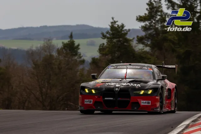 BMW M4 GT3 Nummer 77 in schwarz-roter YOKOHAMA-Lackierung auf der Nürburgring-Nordschleife bei der NLS mit Eifellandschaft im Hintergrund.