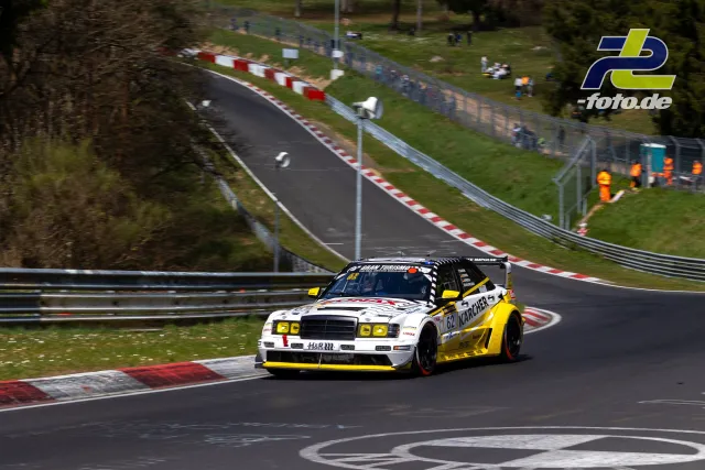 Weißer Renntourenwagen mit gelber Lackierung auf der Nürburgring-Nordschleife bei rotweiß gestreiften Randsteinen.