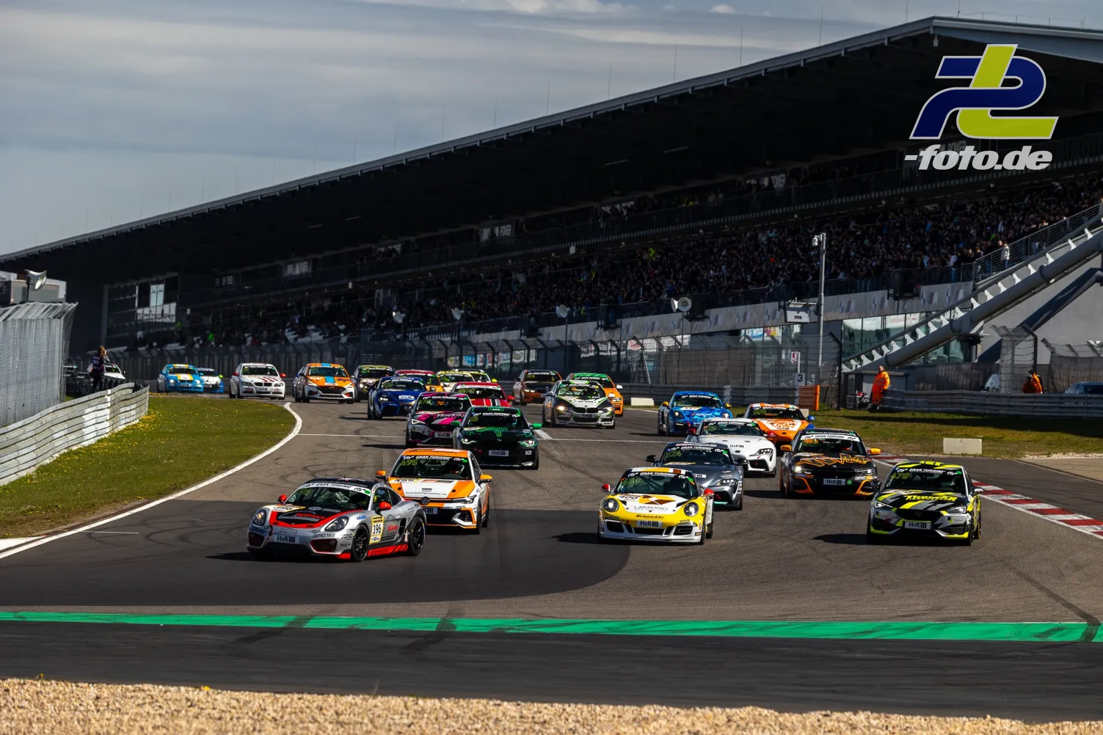 Startphase eines NLS-Rennens auf dem Nürburgring mit großem Fahrerfeld in der ersten Kurve vor gefüllter Haupttribüne.