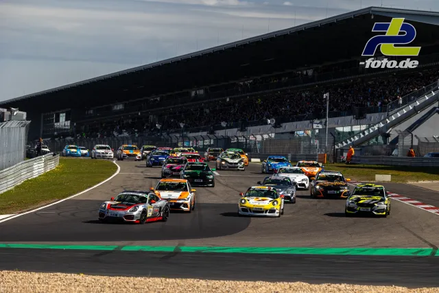 Startphase eines NLS-Rennens auf dem Nürburgring mit großem Fahrerfeld in der ersten Kurve vor gefüllter Haupttribüne.