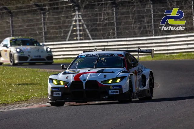BMW M4 GT3 mit weißer Lackierung und roten Akzenten in Kurve auf dem Nürburgring, gefolgt von Porsche