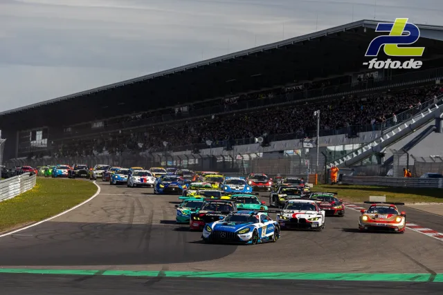 Startphase eines NLS-Rennens am Nürburgring mit dichtem GT3-Feld und voller Haupttribüne im Hintergrund.