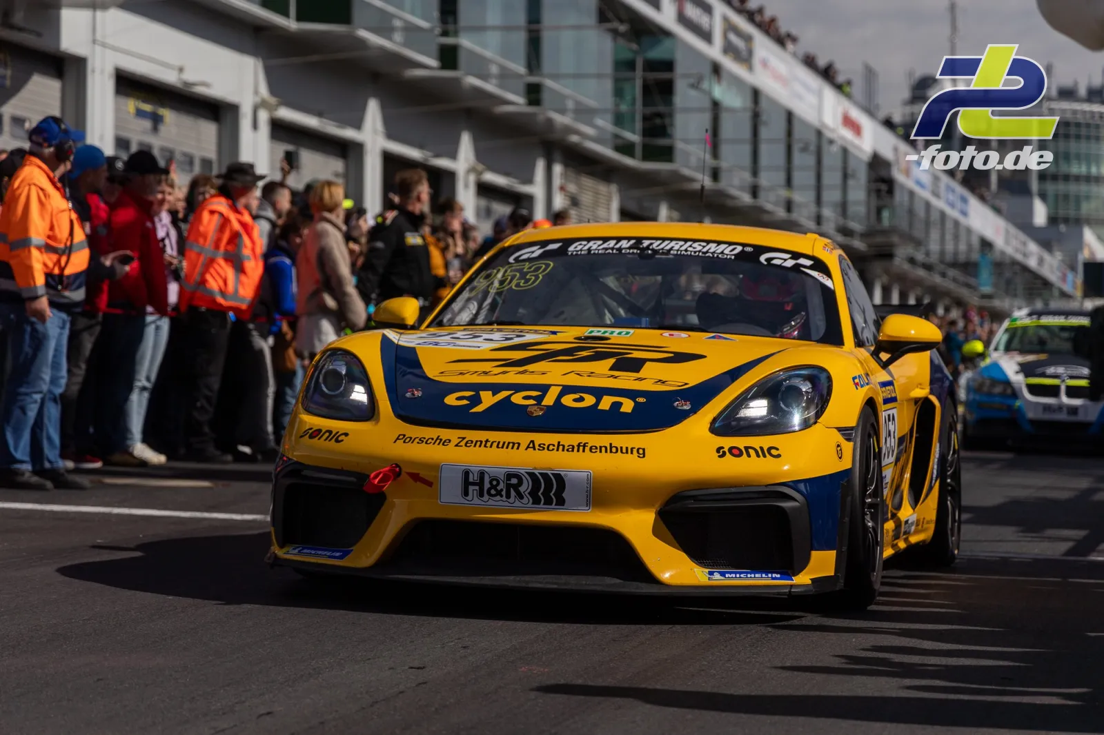 Gelber Porsche 718 Cayman GT4 mit cyclon-Lackierung fährt durch die Boxengasse am Nürburgring, Zuschauer und Streckenposten am Rand.