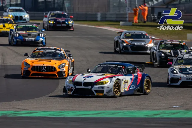 Mehrere GT-Fahrzeuge in einer Kurve auf dem Nürburgring, angeführt von einem BMW Z4 GT3 in Weiß-Rot-Blau und einem orangefarbenen Mercedes-AMG GT.