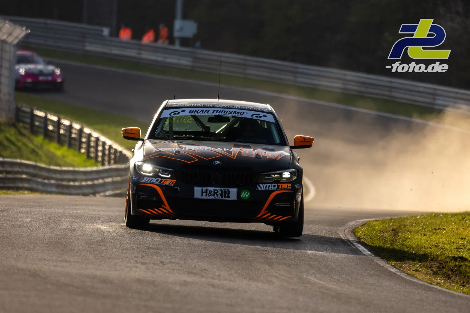BMW M4 GT4 in schwarz-oranger Lackierung auf der Nordschleife, im Hintergrund ein weiteres Fahrzeug, Grünstreifen rechts