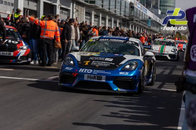 Porsche Cayman mit blauer Lackierung in der Boxengasse des Nürburgrings, umgeben von Team und Zuschauern während der NLS.
