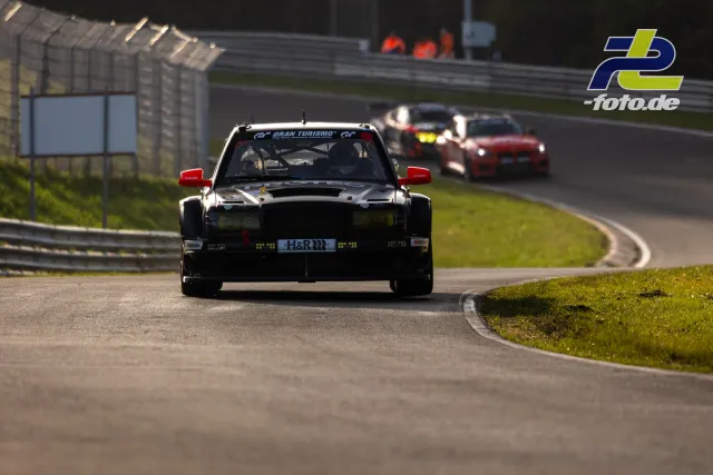 Schwarz-weißer Rennwagen mit roten Spiegeln auf der Nordschleife, gefolgt von mehreren Fahrzeugen durch die Kurve.