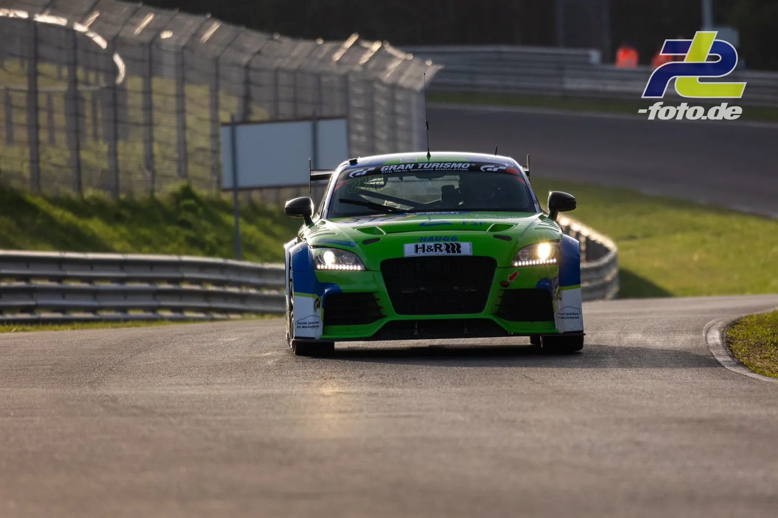 Grün-blau lackierter Rennwagen mit Heckflügel auf der Nordschleife, Frontalansicht während der Fahrt mit eingeschalteten Scheinwerfern.