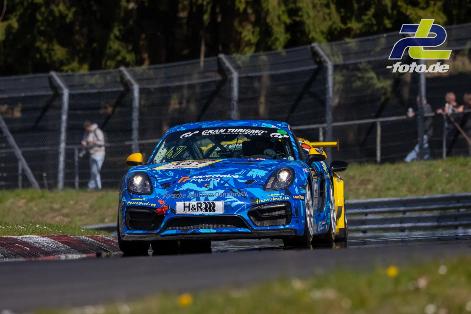 Blauer Porsche mit Startnummer 147 in der Kurve auf dem Nürburgring, gelbes Fahrzeug im Hintergrund, Zuschauer am Streckenzaun.