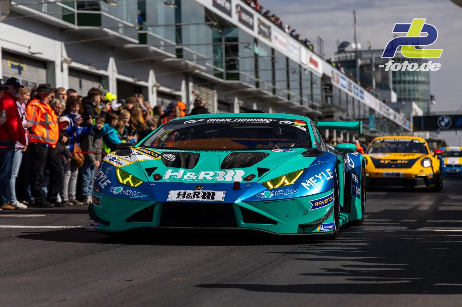 Lamborghini Huracán GT3 in türkis-blau auf der Boxengasse am Nürburgring, gefolgt von gelbem GT3-Fahrzeug, Zuschauer am Boxengebäude.