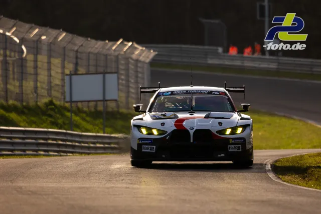 BMW M4 GT3 in weiß-roter Lackierung auf der Nordschleife beim Zeittraining, frontale Ansicht mit leuchtenden Scheinwerfern.