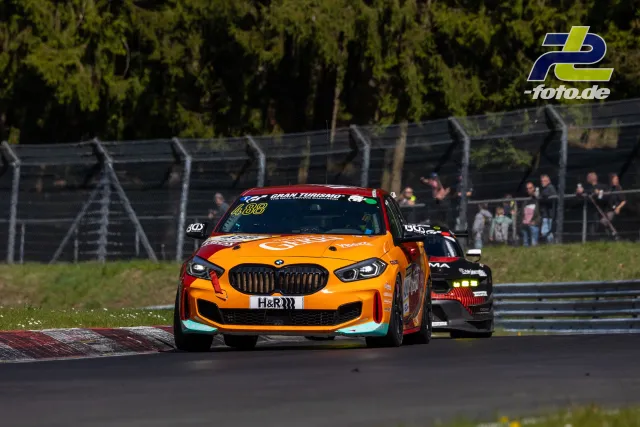 BMW mit orangener Lackierung führt ein schwarzes Fahrzeug in einer Kurve am Nürburgring an, Zuschauer am Streckenzaun.