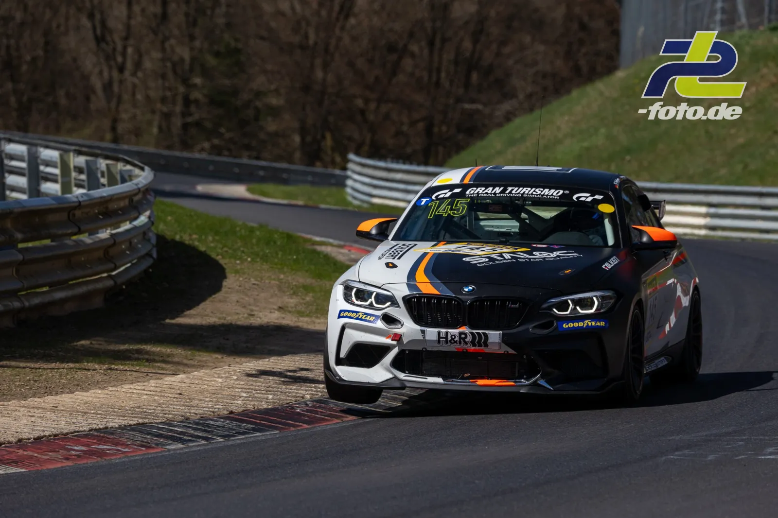 BMW in Kurve auf der Nürburgring-Nordschleife, Startnummer 145, weiße und schwarze Lackierung mit orangefarbenen Akzenten.