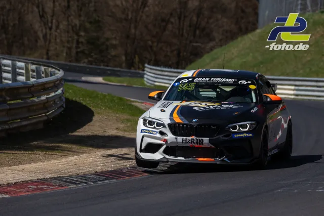 BMW in Kurve auf der Nürburgring-Nordschleife, Startnummer 145, weiße und schwarze Lackierung mit orangefarbenen Akzenten.