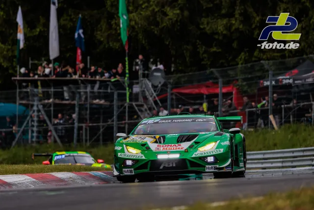 Grüner Lamborghini Huracán GT3 mit eingeschaltetem Licht auf der Nordschleife, im Hintergrund Zuschauertribüne und Fahrerlager