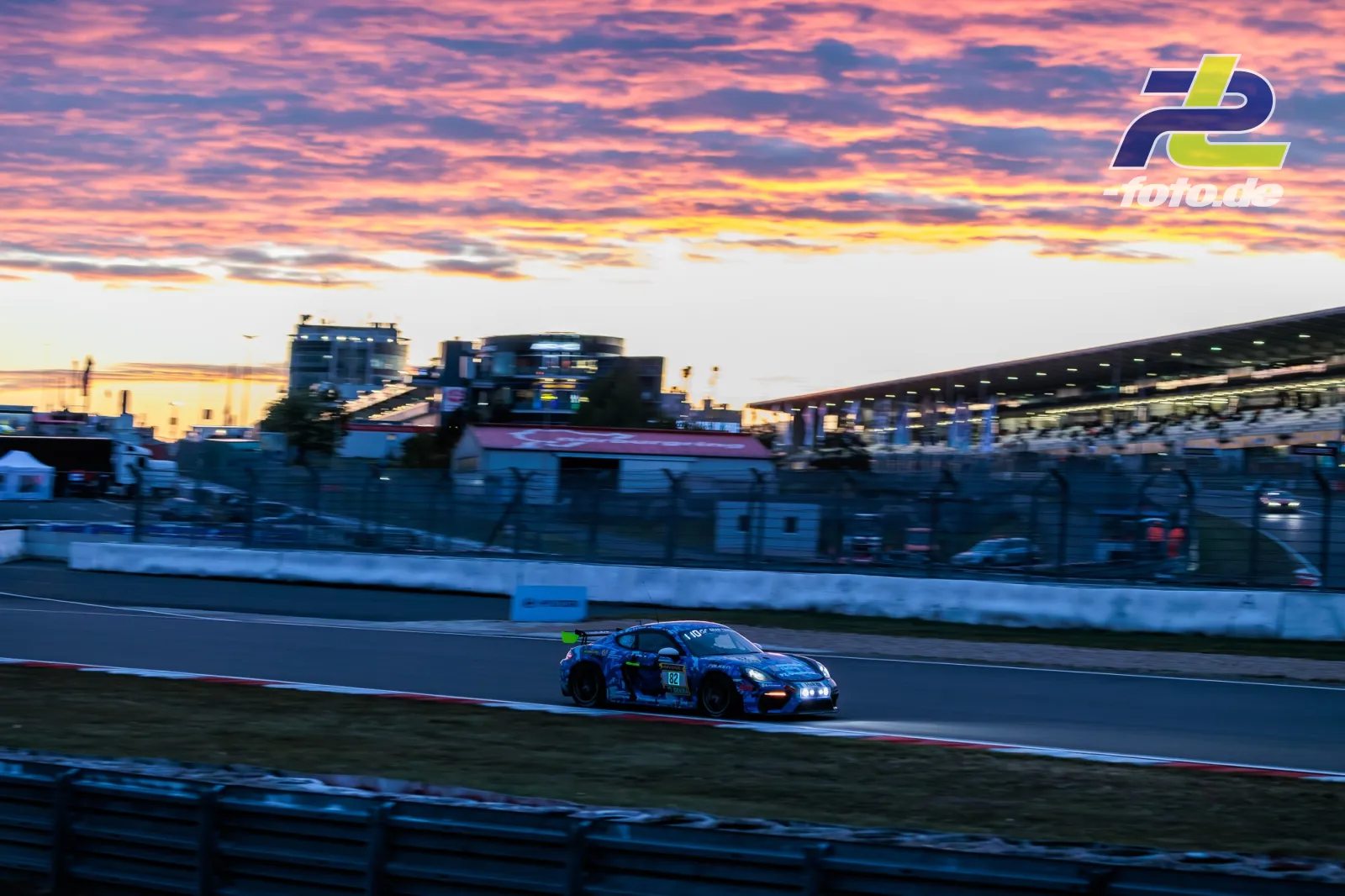 Porsche 718 Cayman GT4 Clubsport beim 24h-Rennen am Nürburgring bei Sonnenuntergang mit dramatischem Abendhimmel und Tribüne im Hintergrund.