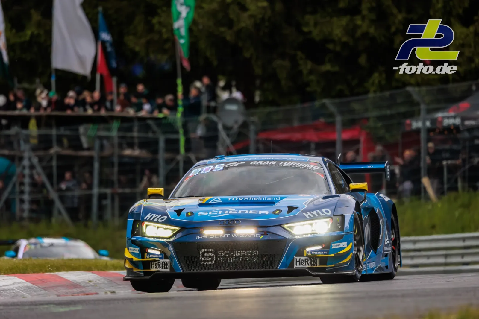Blauer Audi R8 LMS GT3 mit gelben Akzenten während des 24h-Rennens Nürburgring bei Tageslicht vor Zuschauertribüne.