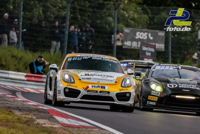 Zwei Rennwagen auf der Nordschleife vor Zuschauern, gelb-weißer Porsche führt vor schwarzem GT3-Fahrzeug.